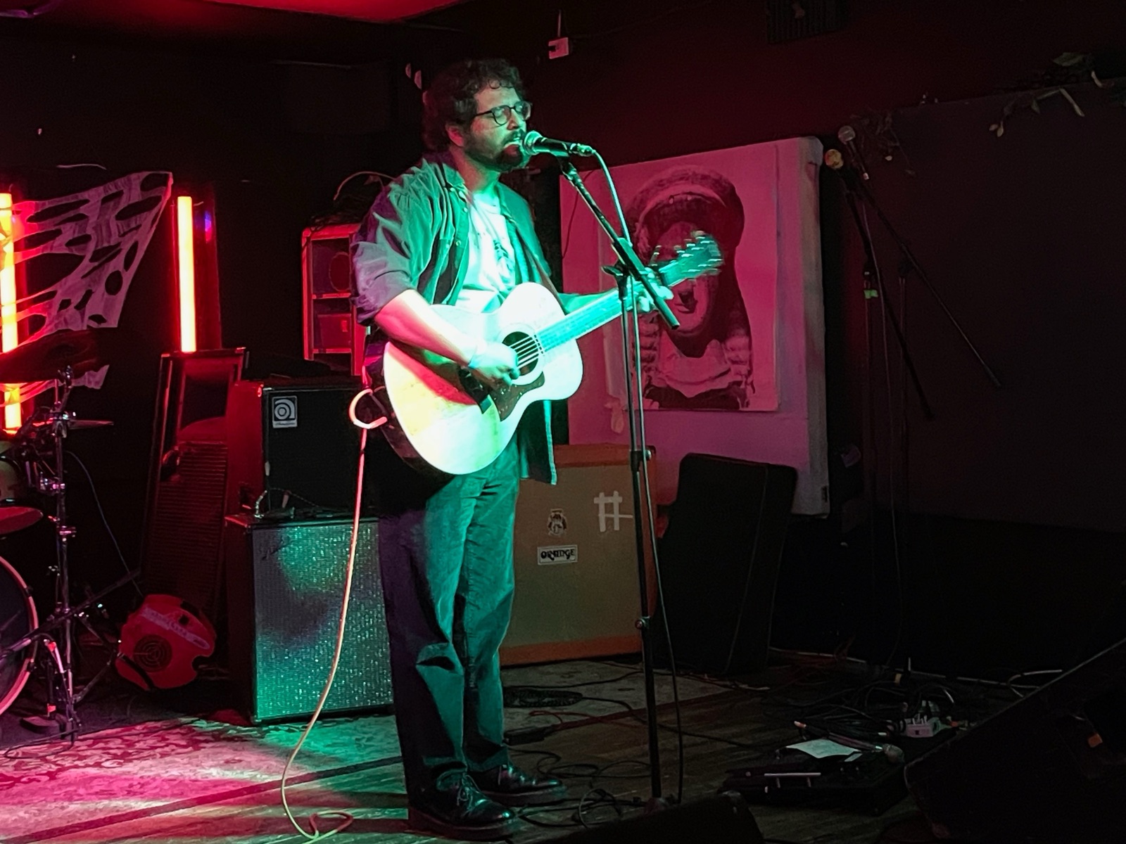 Alex Beckman singing at a mic stand with an acoustic guitar, under red and green stage light.