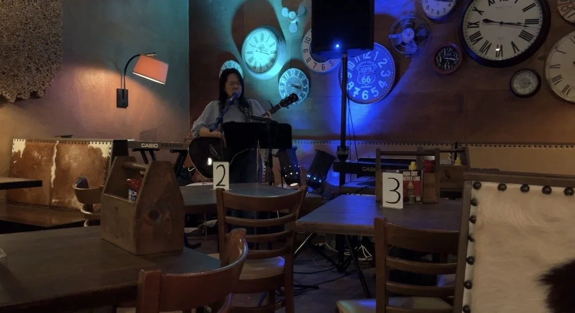 A singer at a microphone with a guitar and two keyboards, under the clocks on the back wall at 317 Burger.