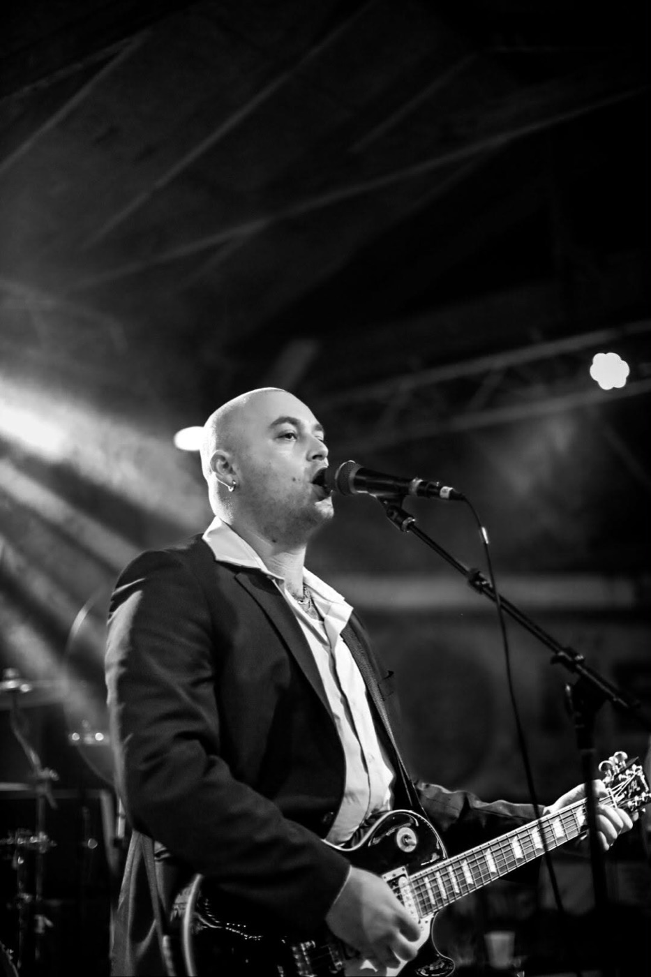Muddy Marshall singing into a microphone with an electric guitar, in black and white under stage lights.