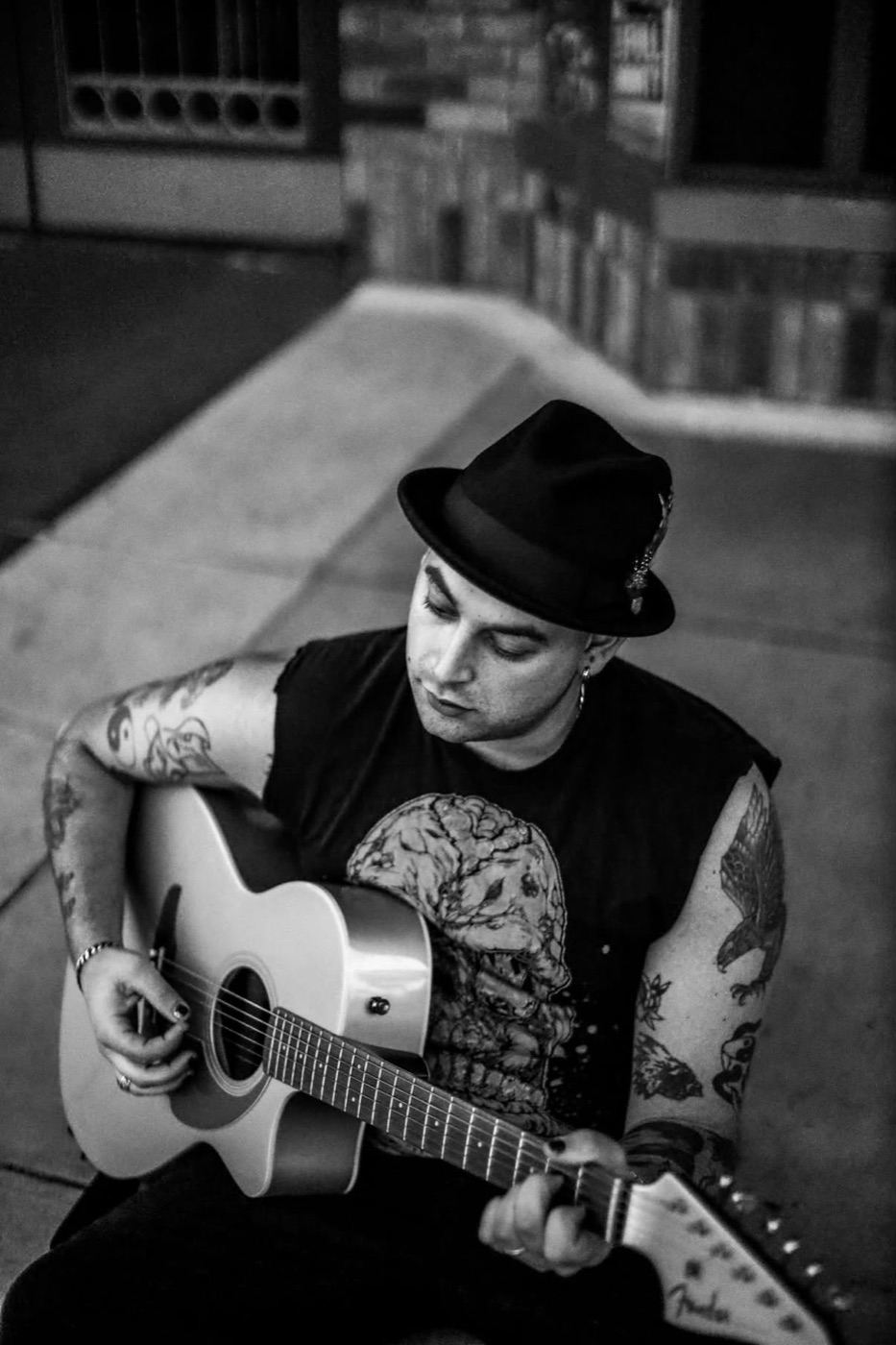 Muddy Marshall in a hat, playing an acoustic guitar on a sidewalk, in black and white.