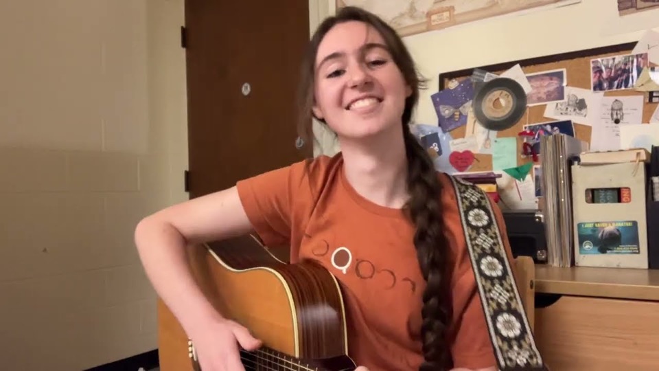 Pocket Diver smiling with an acoustic guitar in a dorm room, a corkboard of postcards and a 45 record behind her.