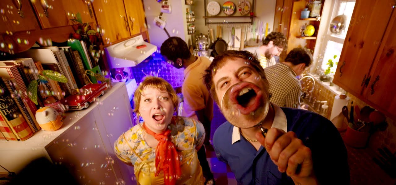 Two members of Wishy singing toward the camera through soap bubbles in a kitchen, one holding a magnifying glass over his mouth.